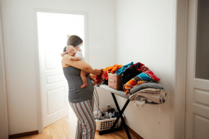 Parent tenant un bébé près d’une pile de linge, montrant un espace de vie saturé et mal organisé