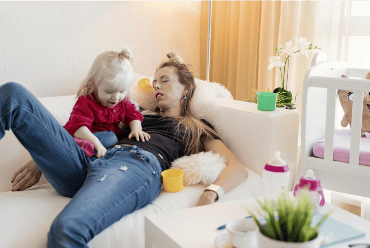 Parent allongé sur un canapé avec un enfant, illustrant la fatigue du quotidien dans un intérieur encombré