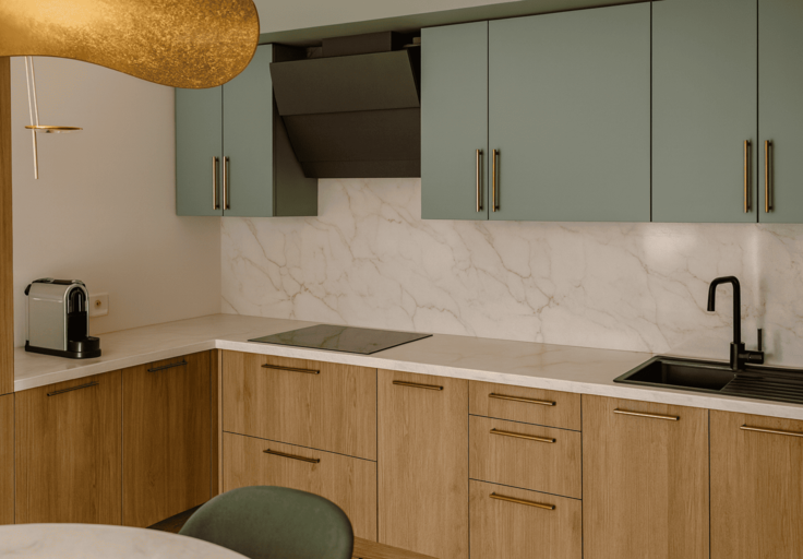 Cuisine en L avec façades bois et vert sauge, hotte intégrée, crédence marbre — aménagement Studio Intéa.